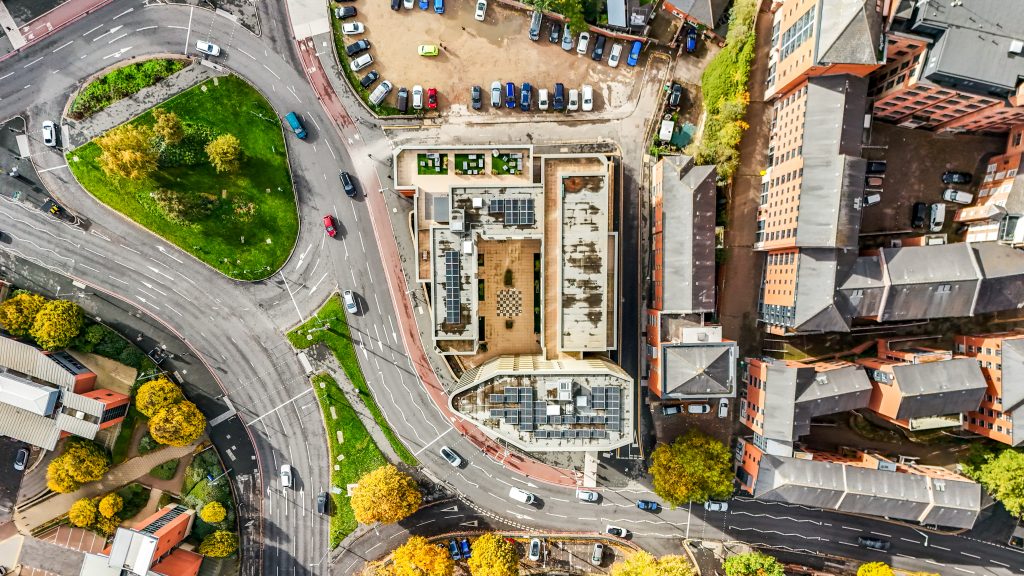 Aerial view of a city intersection with a building featuring a rooftop garden and surrounding vehicles parked along the streets.