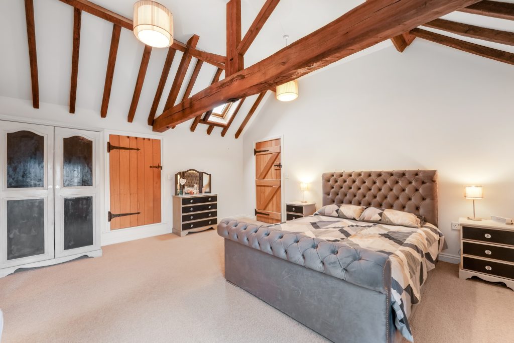 Bedroom with a tufted gray bed, wooden beams on the ceiling, and matching nightstands with lamps.