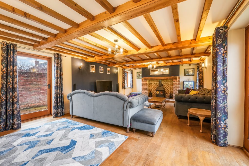 Living room with a blue sofa, wooden beams, and a brick fireplace, featuring a patterned rug and large windows.