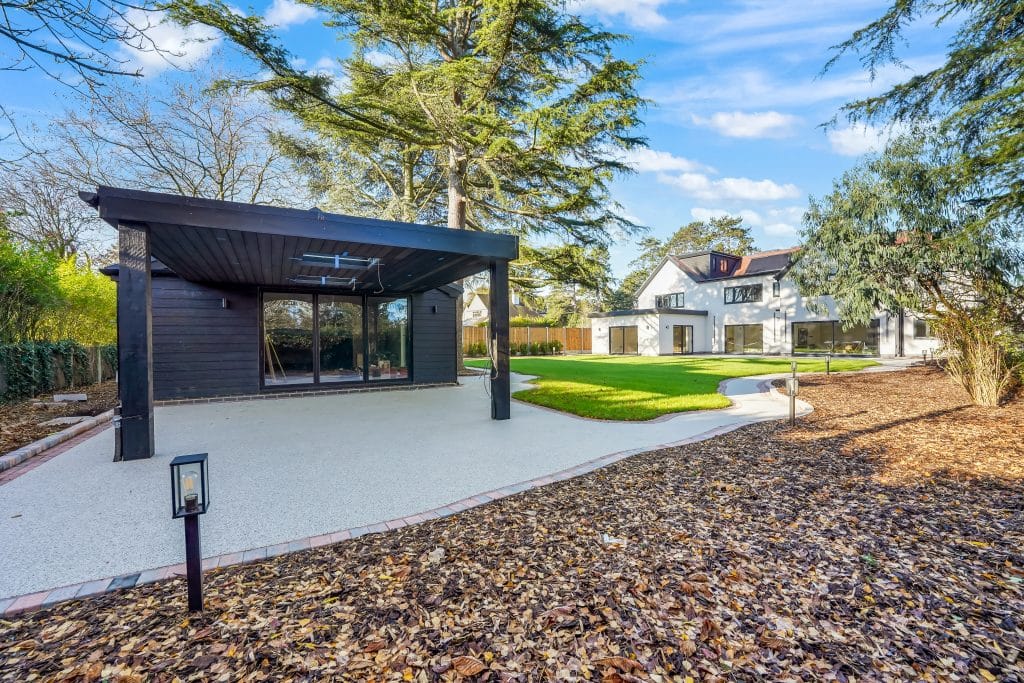 Black wooden structure with large glass windows in a landscaped garden area featuring grass and trees.